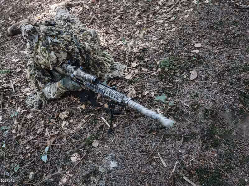 A soldier wearing a ghillie suit is lying on the ground.