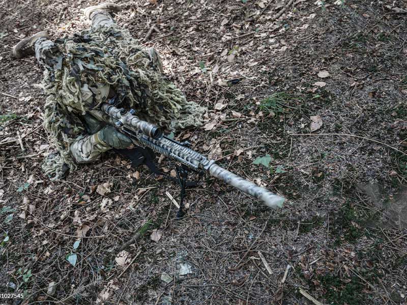 A soldier wearing a ghillie suit is lying on the ground.